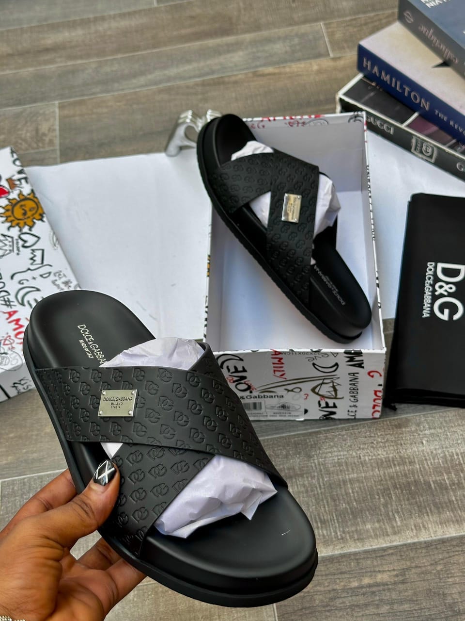 Black Dolce & Gabbana monogram slides with logo plaque held in hand and shown in box with branding.