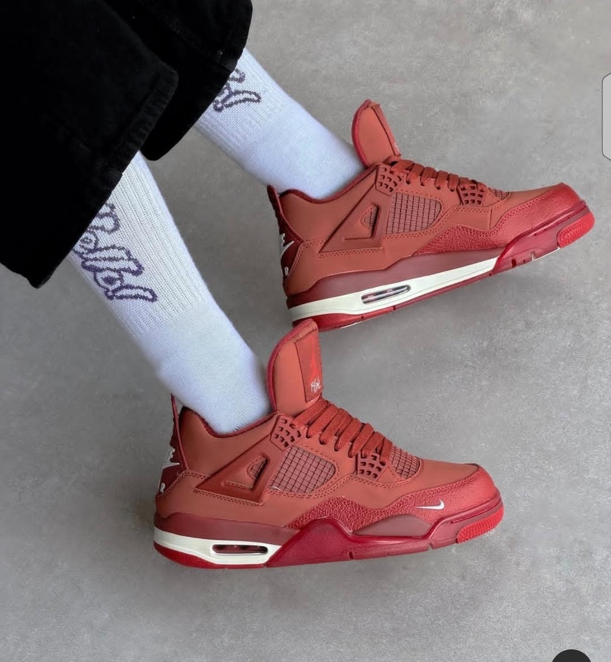 Red Air Jordan 4 sneakers worn on feet with white socks and black pants on grey floor.