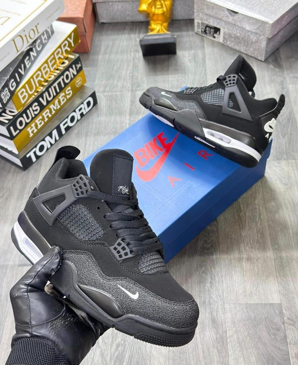 Black Air Jordan 4 sneaker held by gloved hand with blue Nike box and luxury books in background.