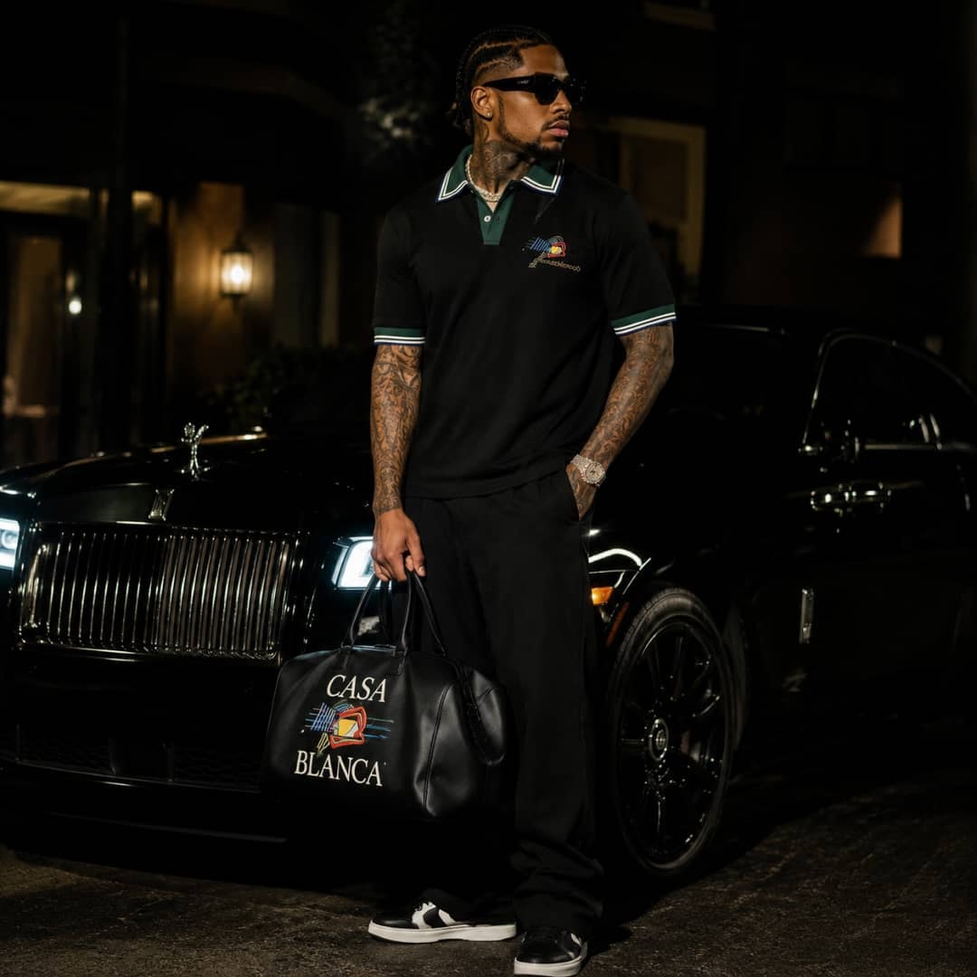 Man wearing black green-trim polo shirt holding bag beside luxury car at night.