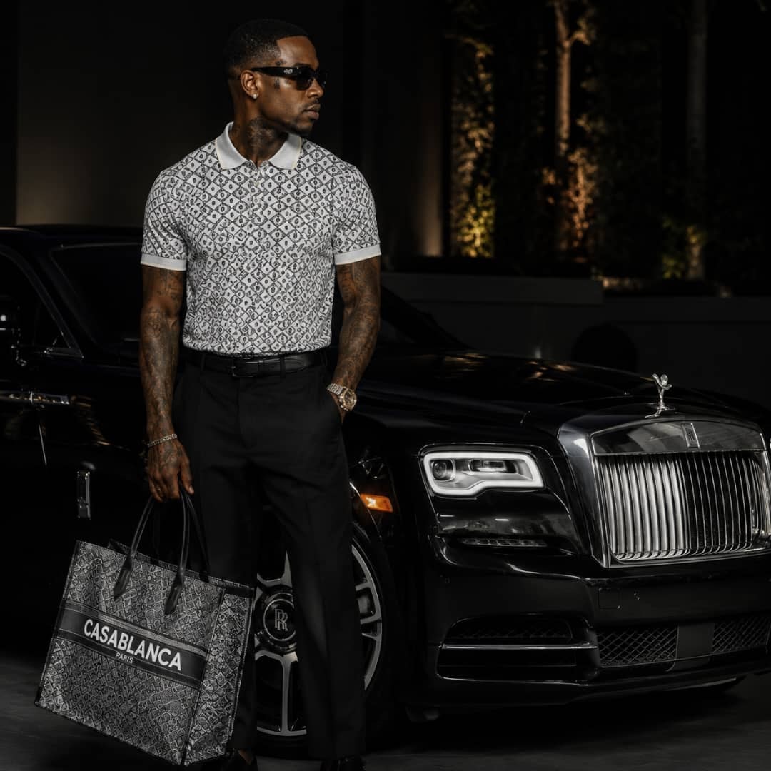 Man wearing black and white patterned polo shirt next to luxury car.
