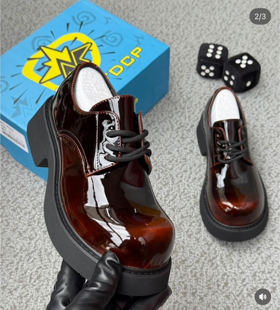 Brown patent leather platform derby shoes held in gloved hand with blue product box and dice in background.