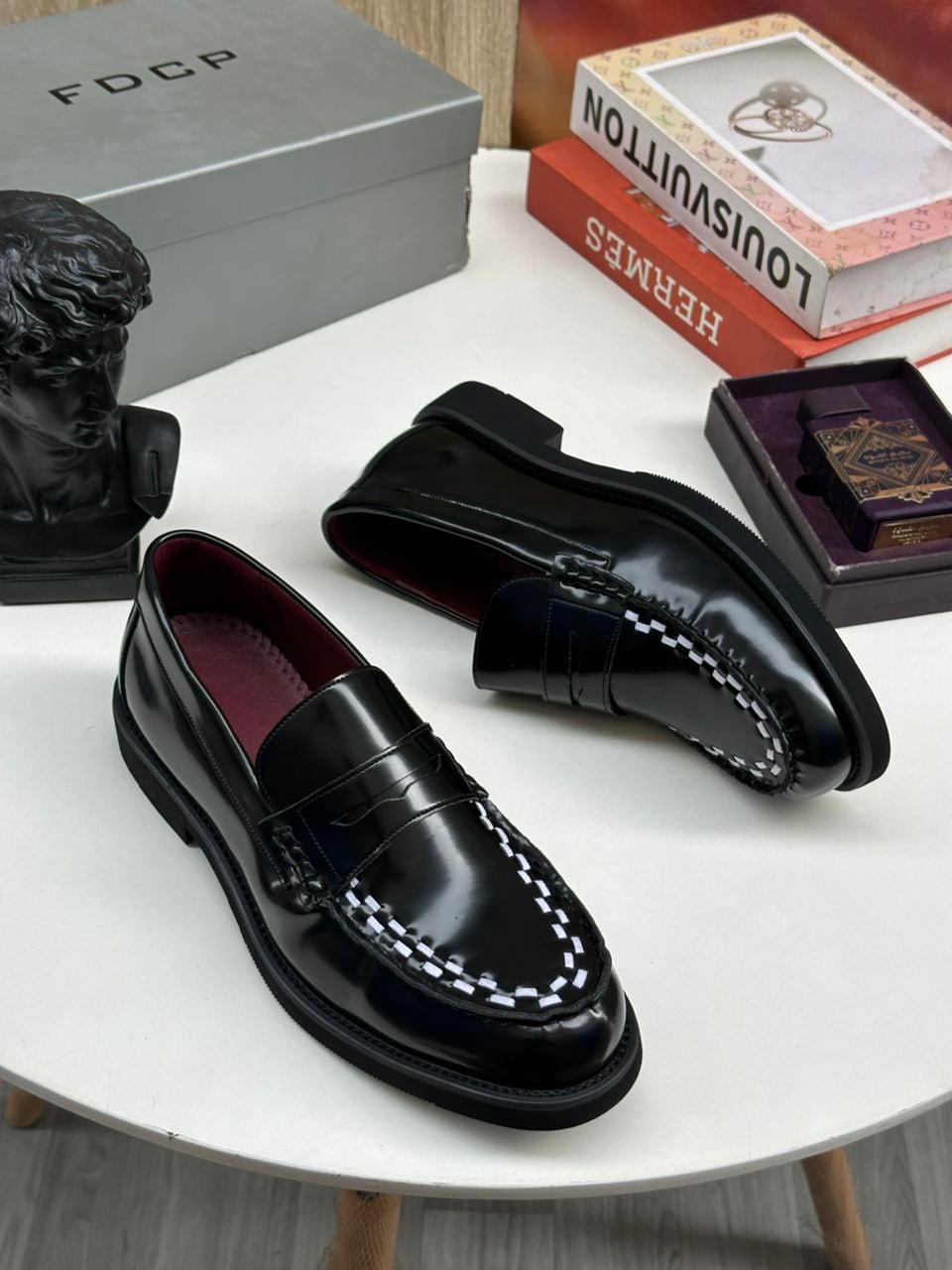 Black patent leather loafers with white stitching on white table with luxury boxes and decor.