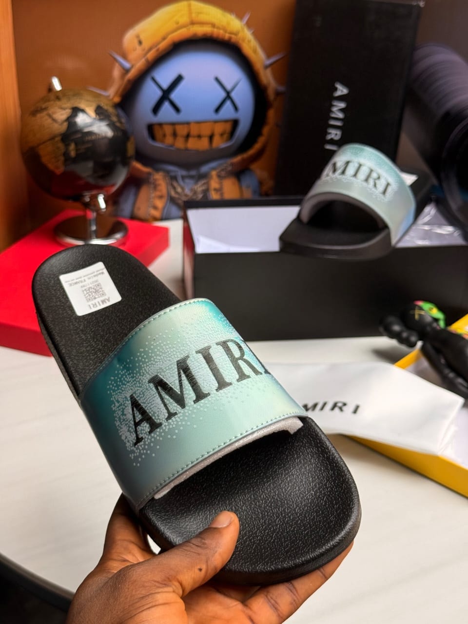 AMIRI slides with teal gradient strap and black sole, held in hand.