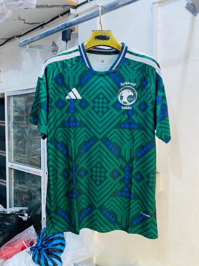 Green geometric patterned Saudi Arabia soccer jersey hanging on yellow hanger.