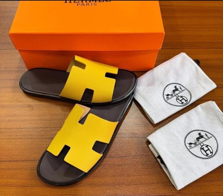Yellow Hermes H-shaped sandals shown with orange box and branded dust bags on wooden surface.