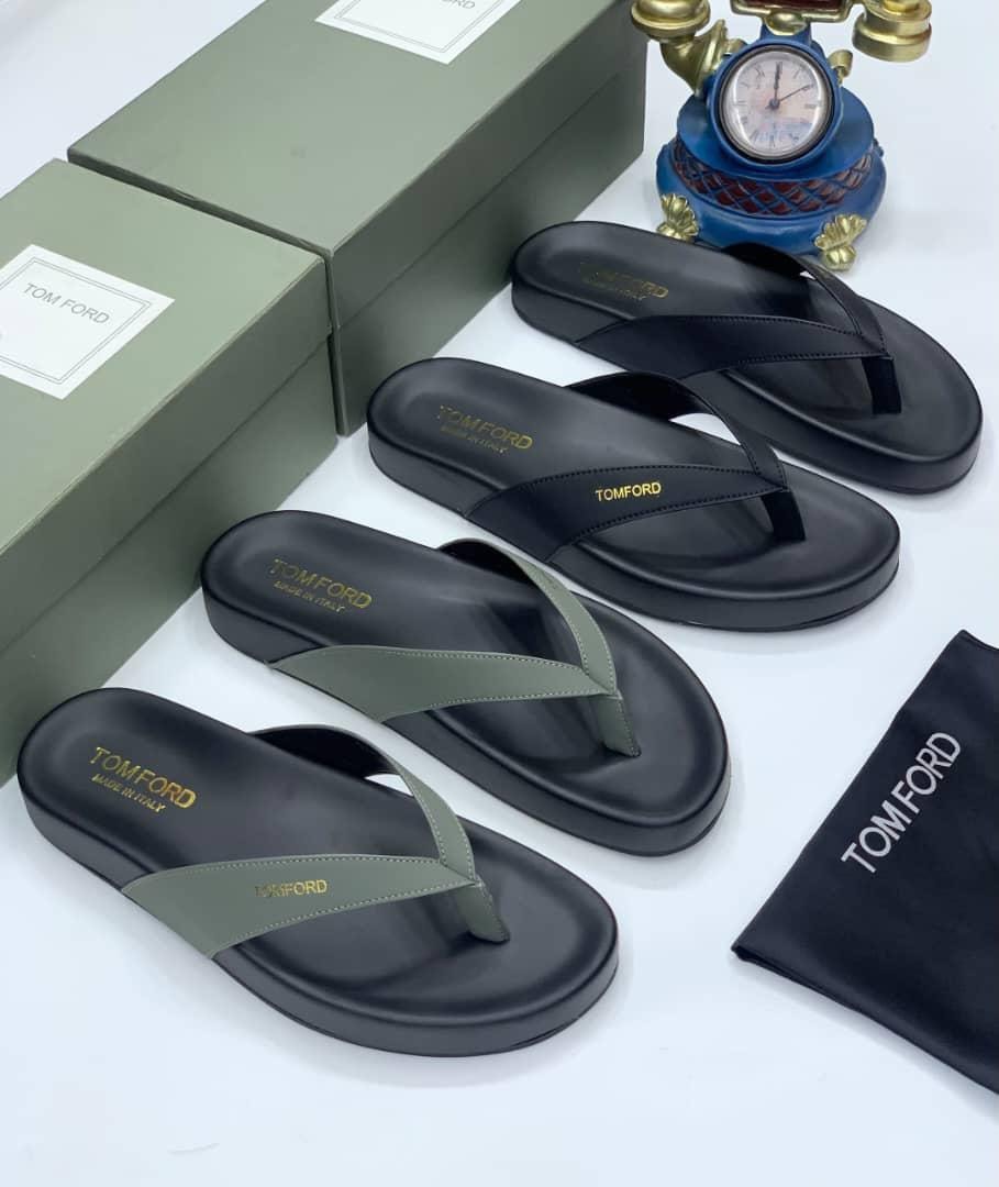 Tom Ford flip flops in black and olive displayed with green boxes, clock and dust bag on white surface.