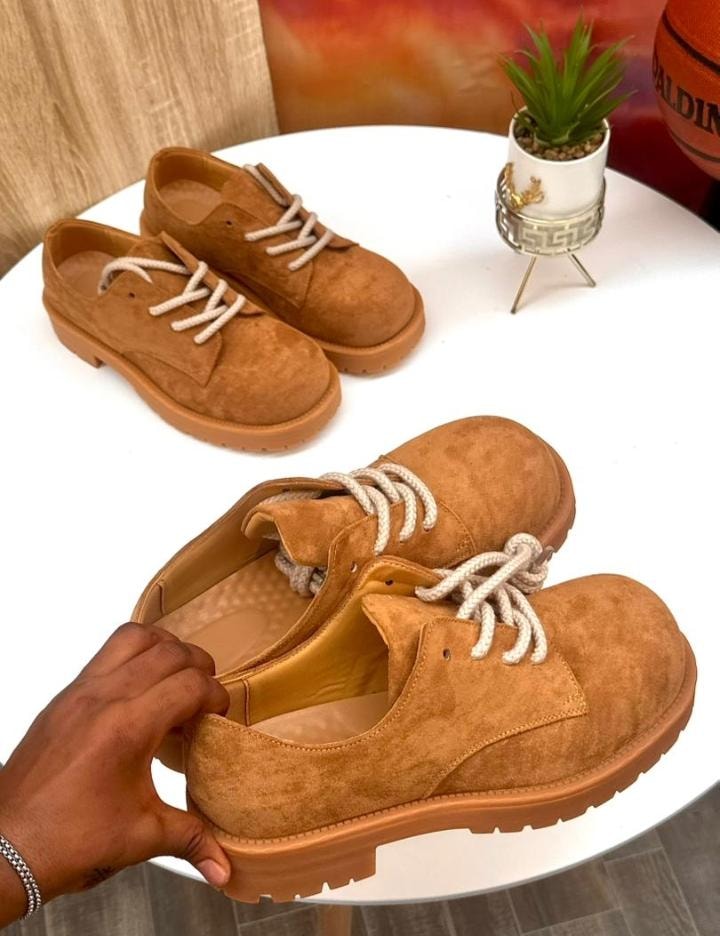 Brown suede lace-up shoes with thick soles displayed on white table, one held by hand showing interior.