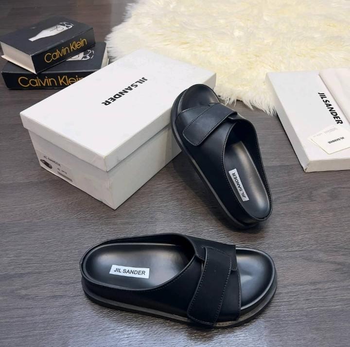 Black Jil Sander single-strap slide displayed with product boxes on gray surface.