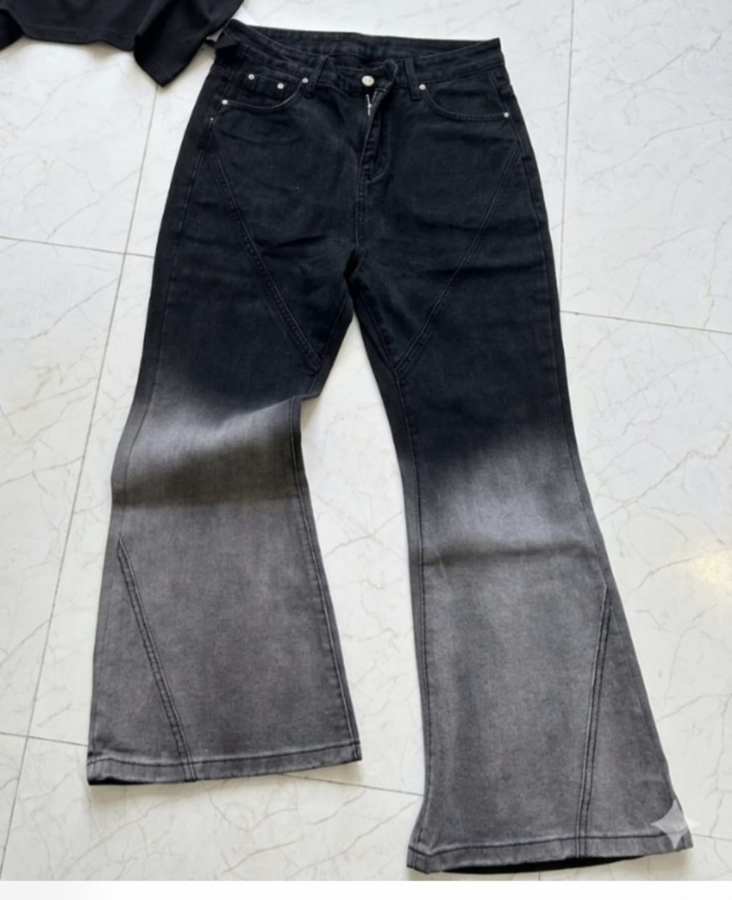 Black to gray ombre flared jeans with gradient wash and seam accents laid flat on tiled floor.