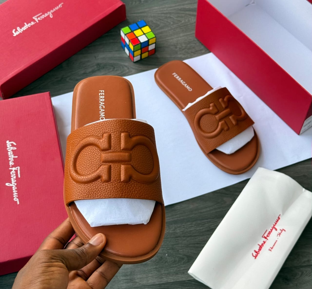 Hand holding brown Salvatore Ferragamo slide with large Gancini logo on strap.