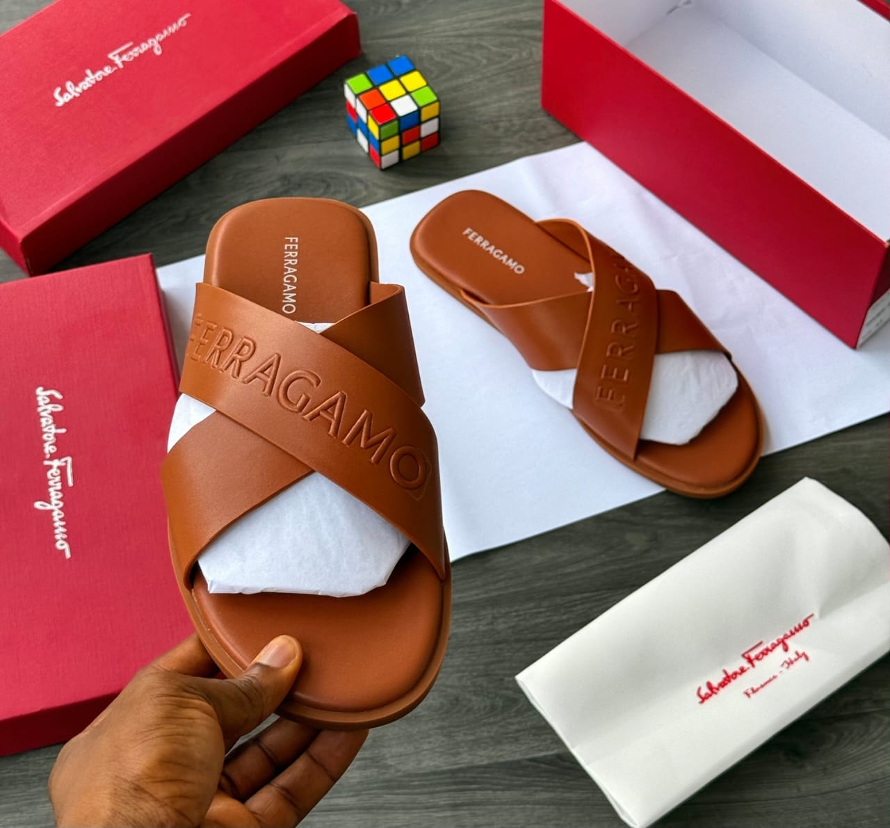 Hand holding brown Salvatore Ferragamo cross strap slide with logo embossing.