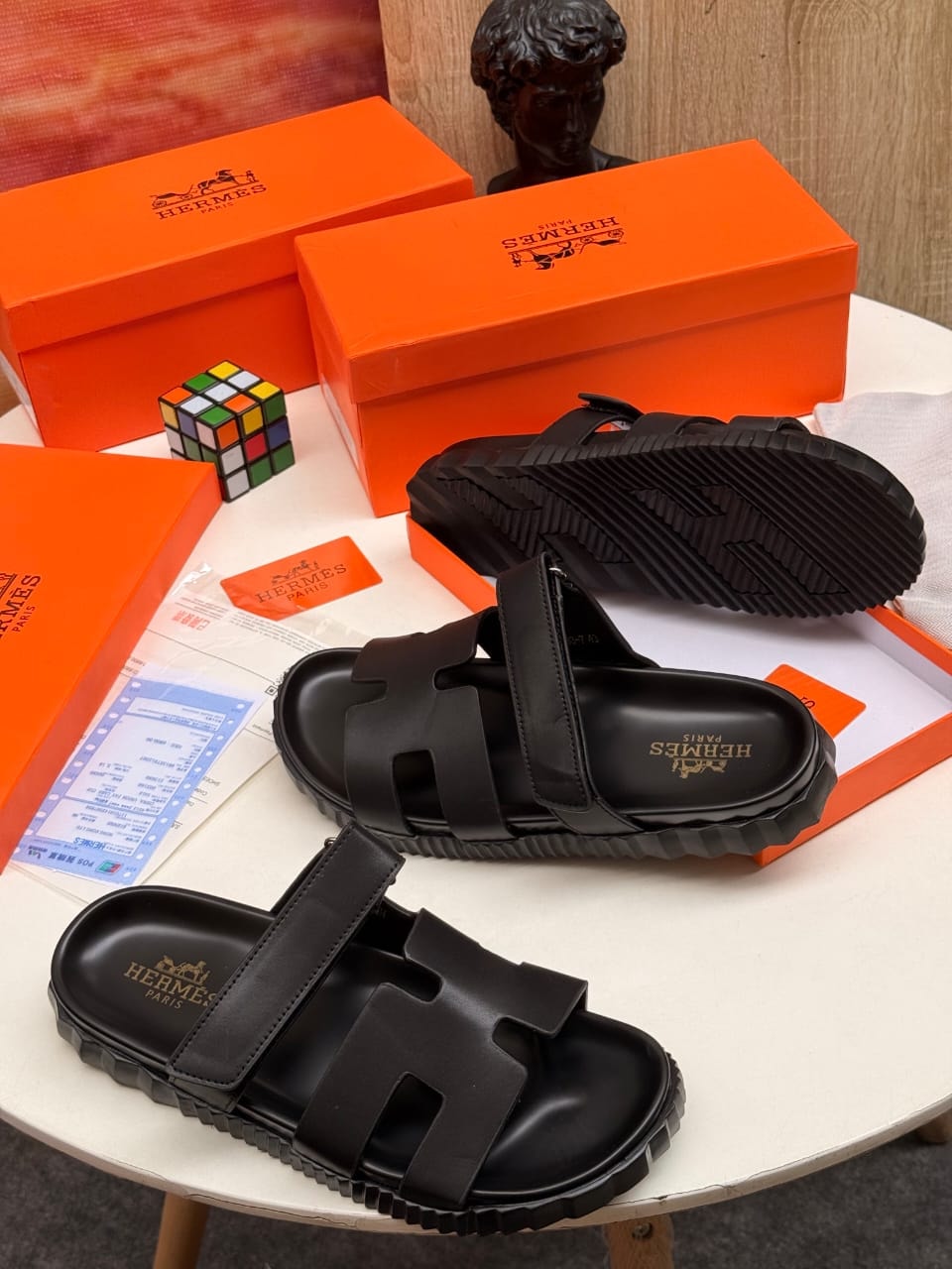Black Hermes chunky H sandals displayed on table with boxes and accessories.
