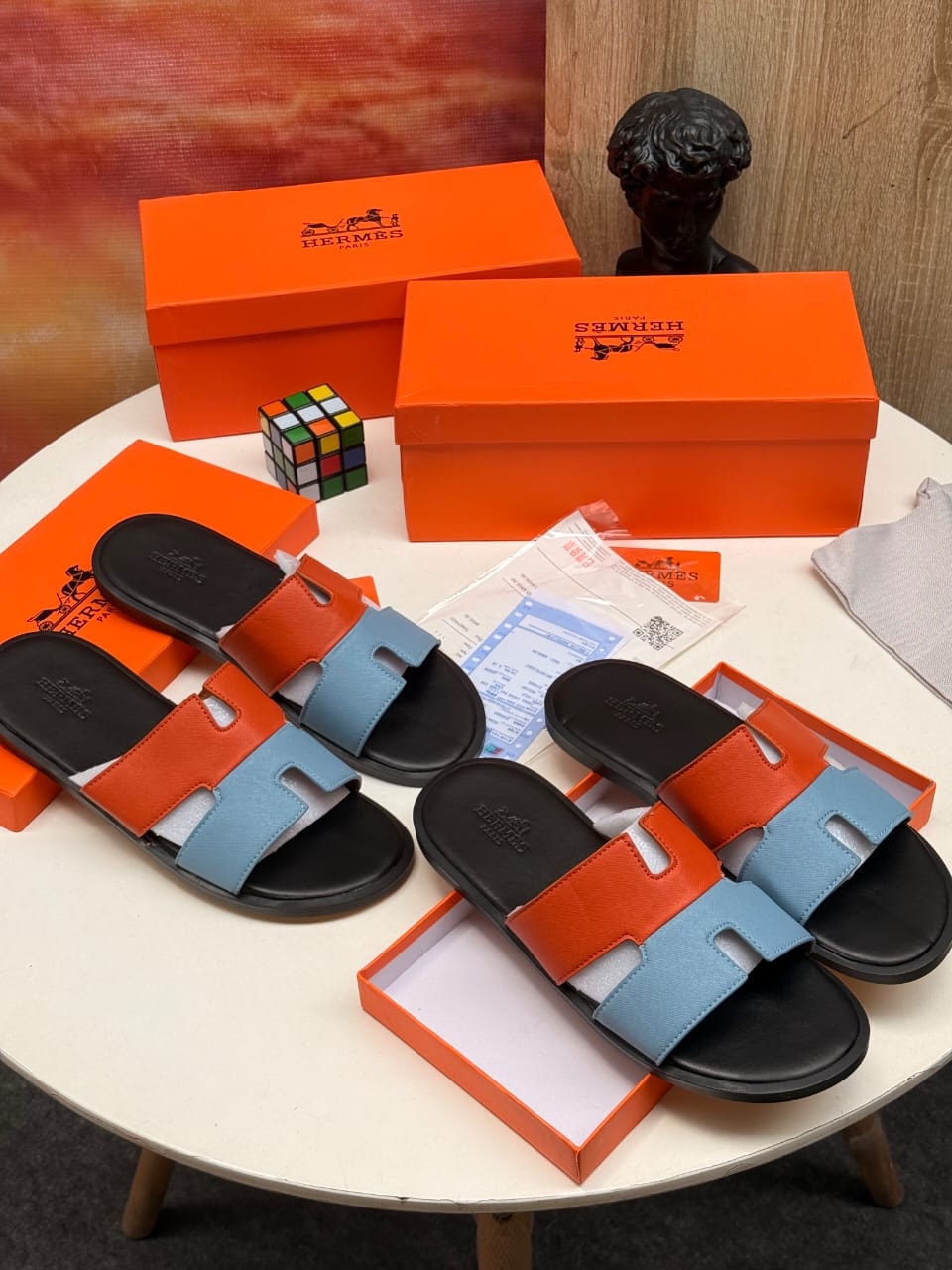 Two pairs of Hermes bi-color H cutout sandals in orange-blue and black-blue with full packaging.
