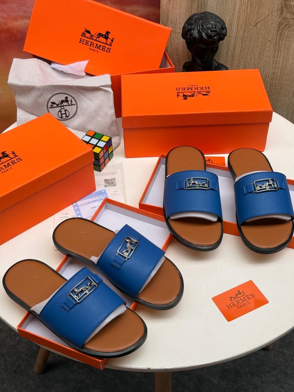 Blue Hermes single strap slides with H buckle and packaging on white table.