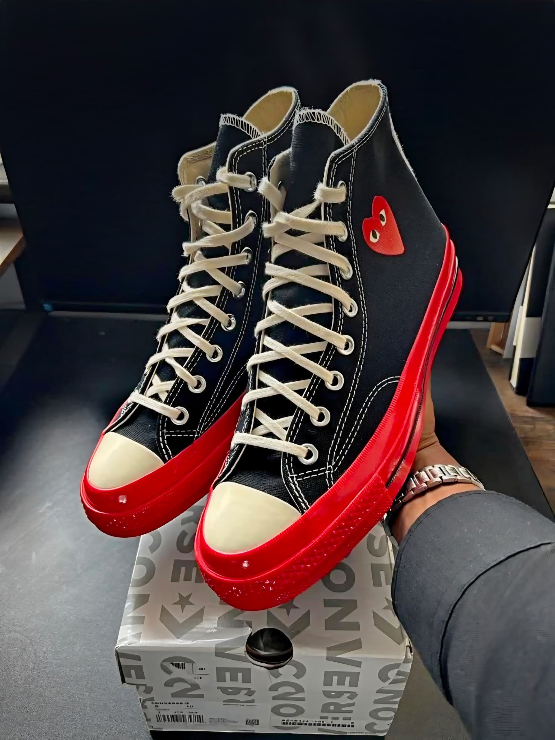 Pair of black high-top Converse sneakers with red soles, white laces, and red heart logo held above the Converse shoebox.