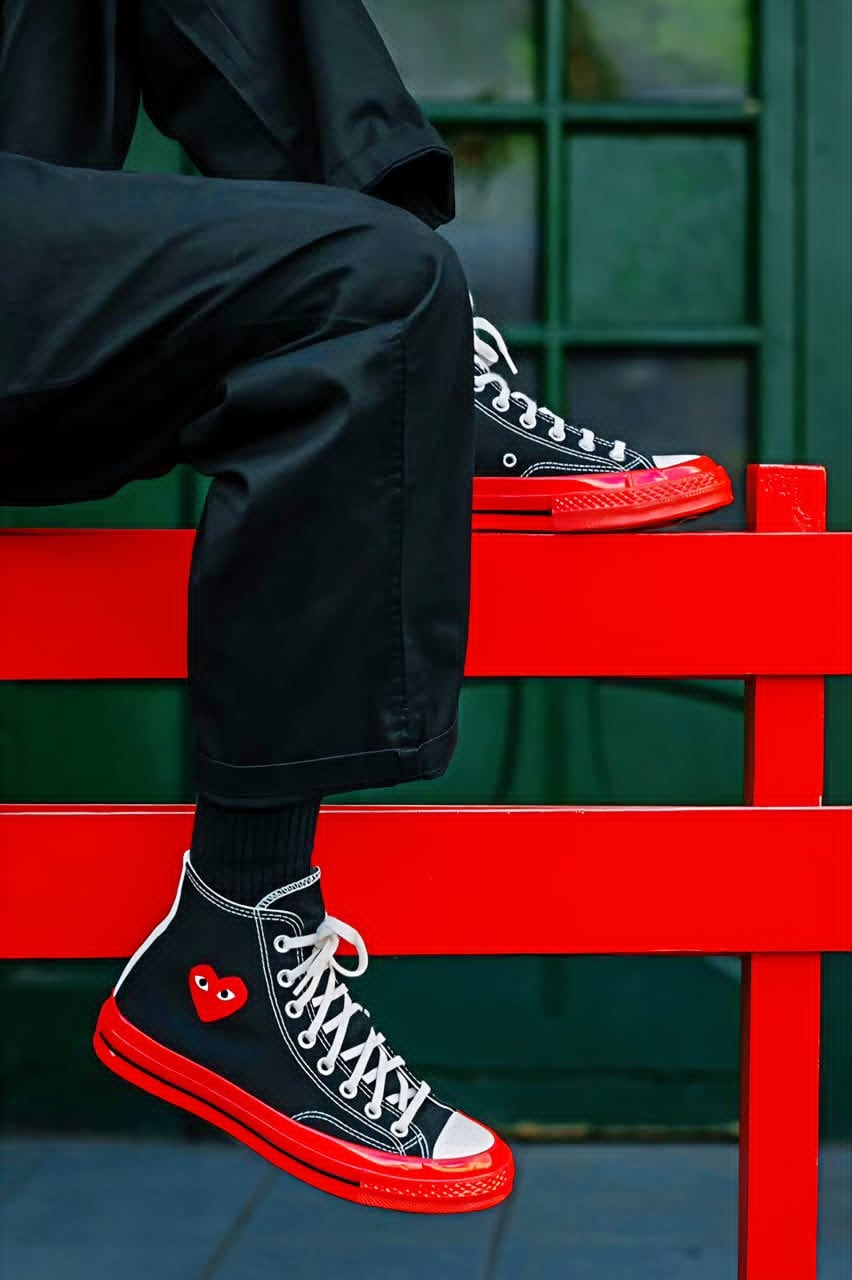 Person wearing black high-top Converse sneakers with red sole and heart patch while seated on a red bench.