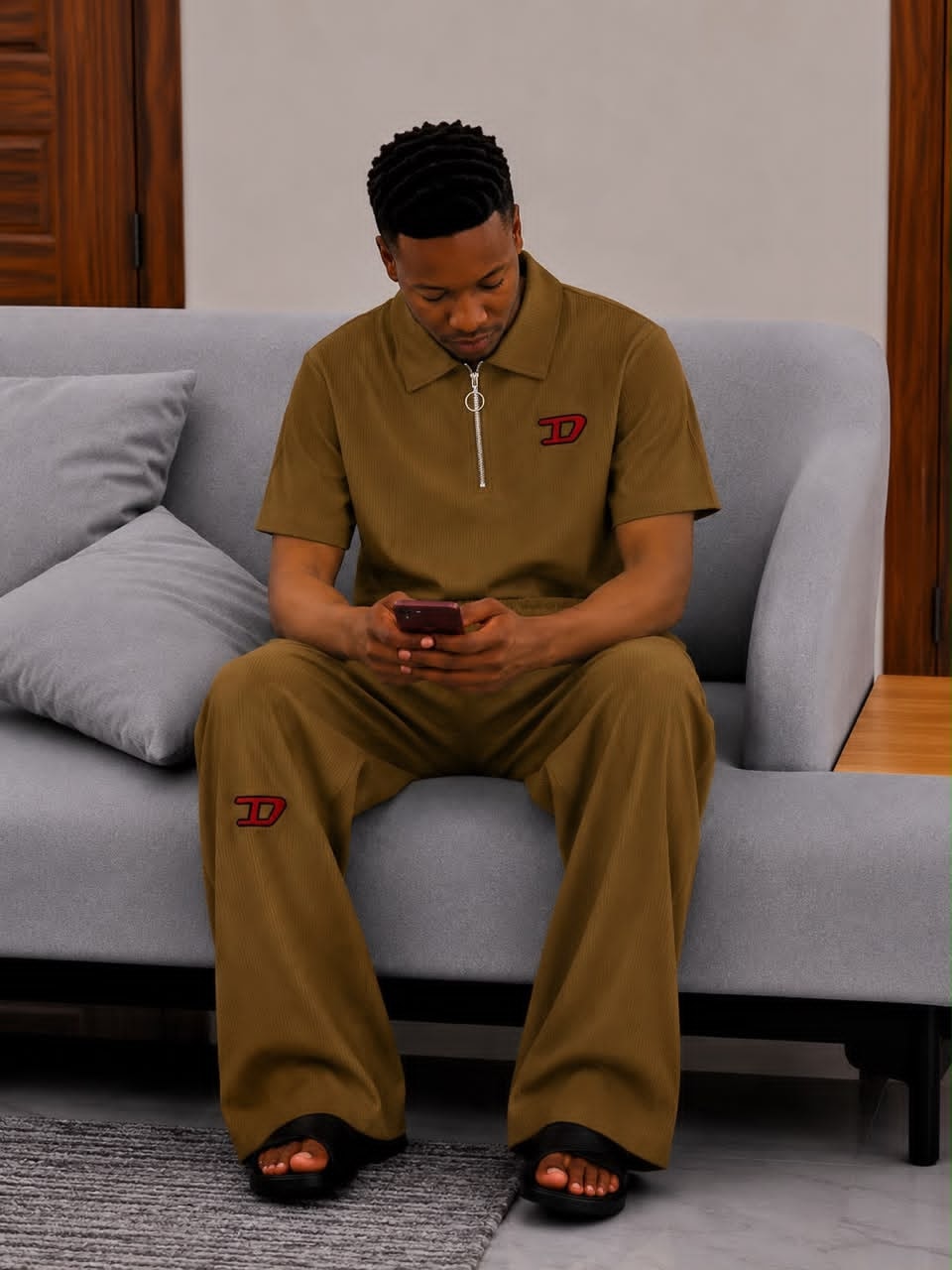 Man seated on gray sofa wearing olive ribbed two-piece set with red D logo on chest and leg.