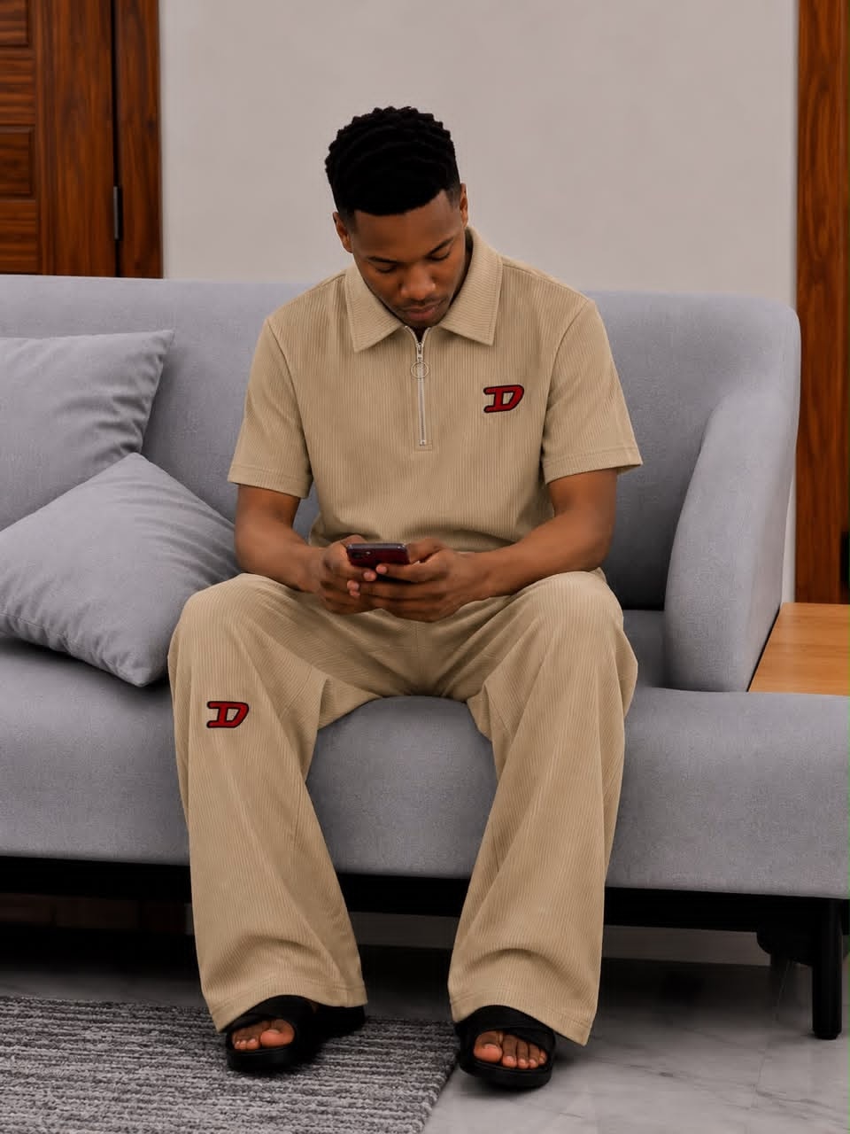 Man seated on gray sofa wearing beige ribbed two-piece set with red D logo on chest and leg.