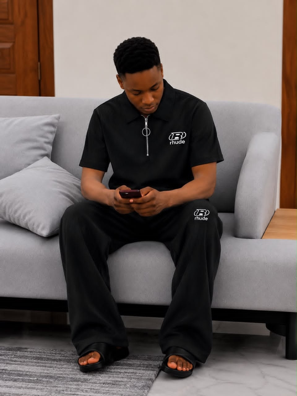 Man seated on gray sofa wearing black ribbed two-piece set with white Rhude logo on chest and leg.