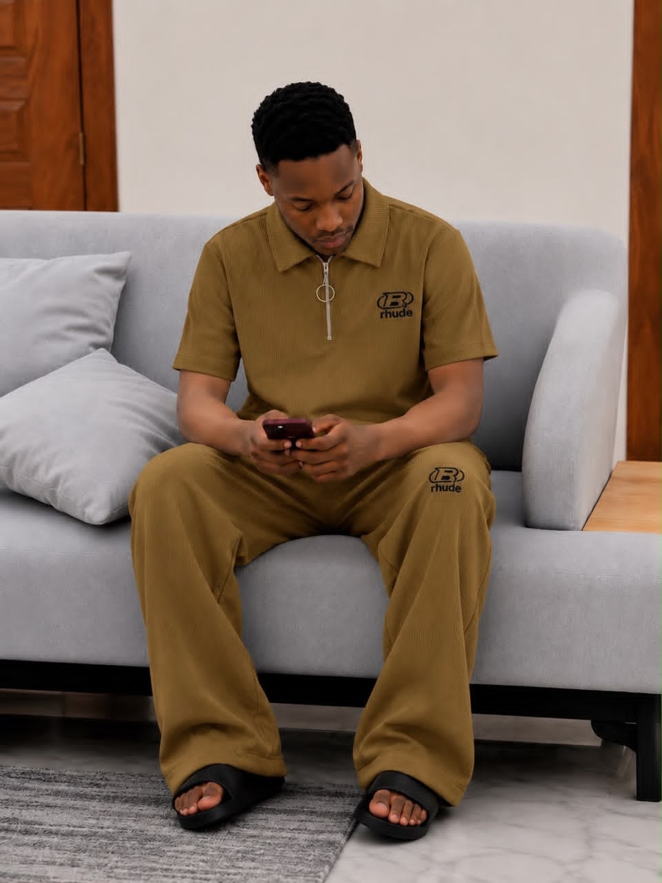 Man seated on gray sofa wearing olive ribbed two-piece set with black Rhude logo on chest and leg.