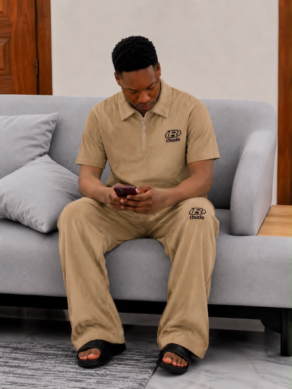 Man seated on gray sofa wearing beige ribbed two-piece set with black Rhude logo on chest and leg.