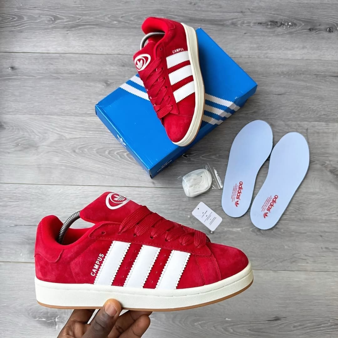 Red Adidas Campus sneakers with white stripes, box, and accessories on wooden surface.