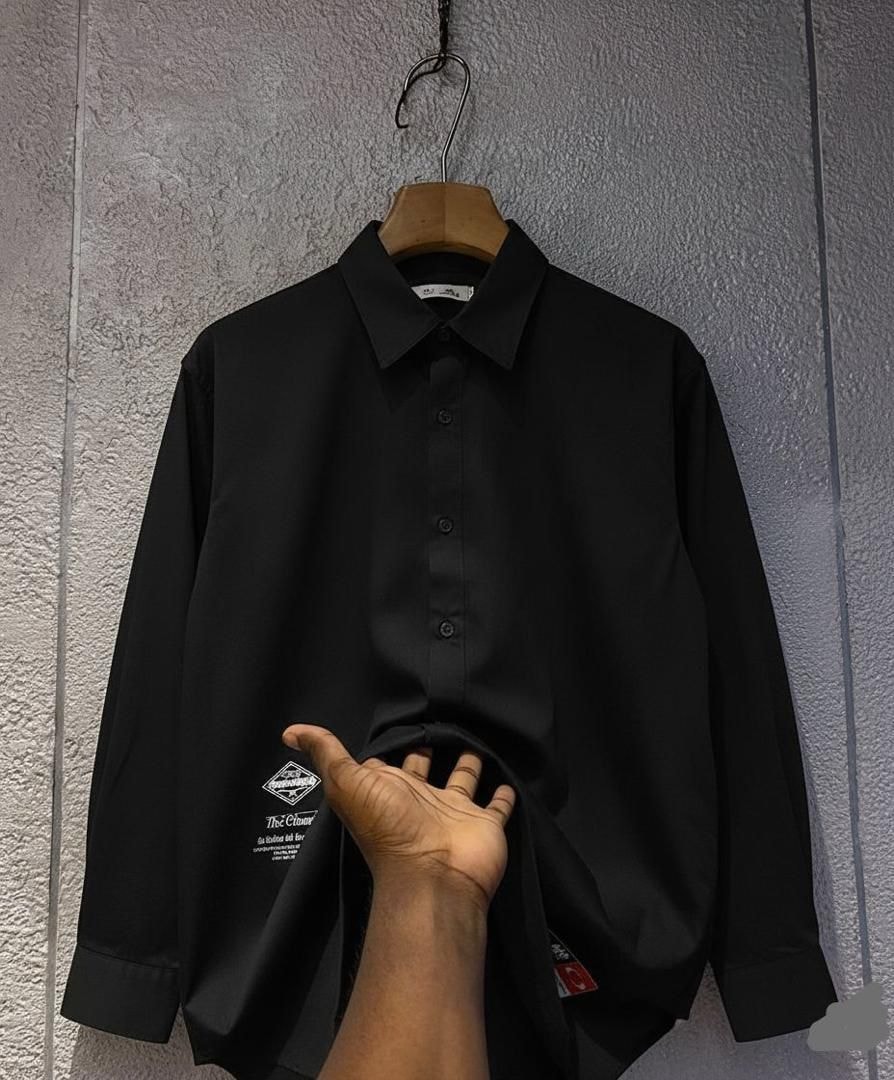 Black long sleeve button-up shirt with chest logo held at the hem against a wall.