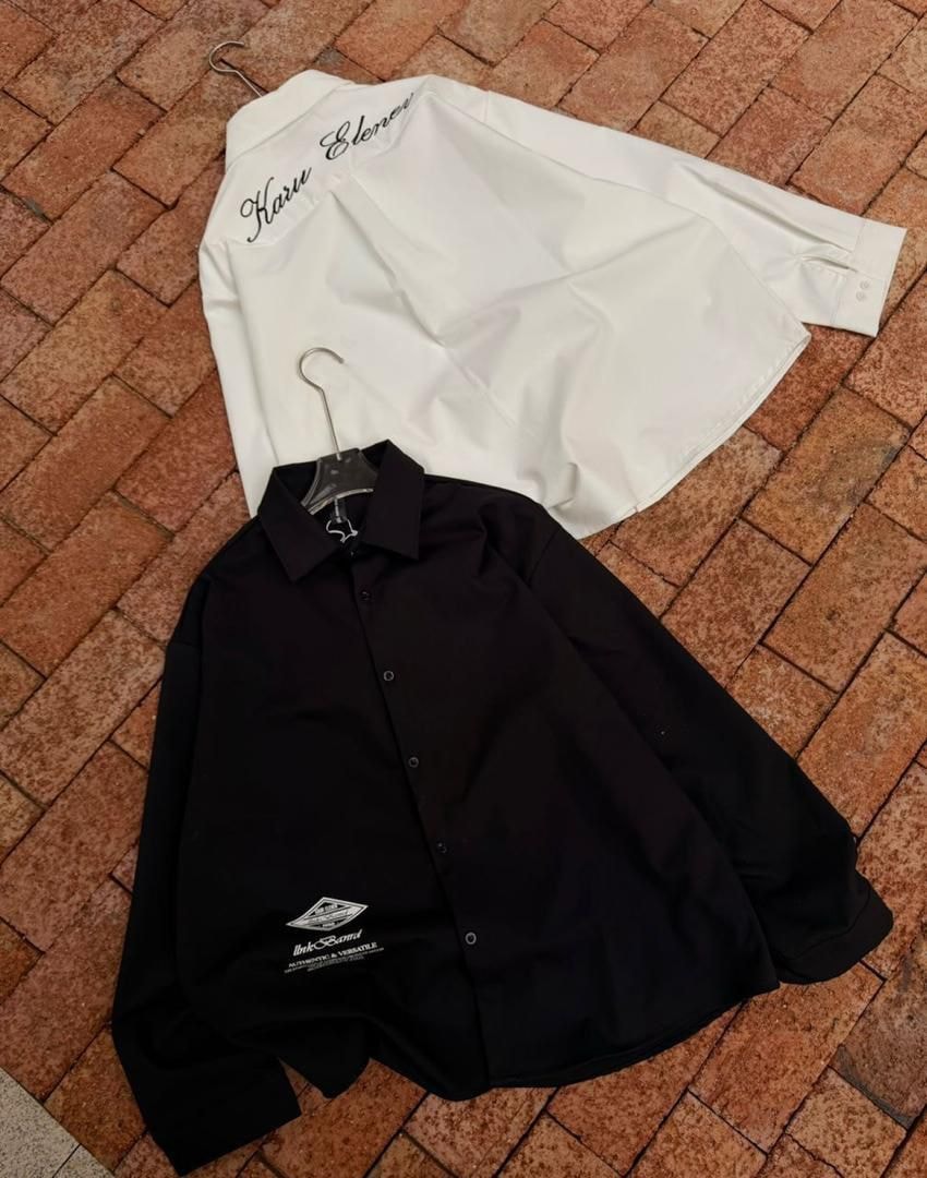 White and black long sleeve button-up shirts on brick pavement showing back embroidery and front logo.