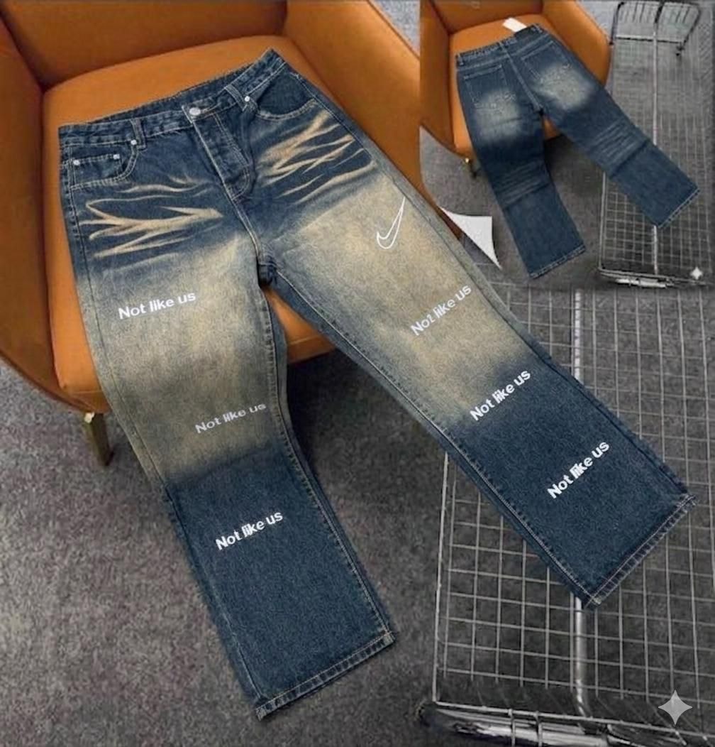 Blue faded baggy jeans with 'Not like us' repeated text and white swoosh logo on chair.