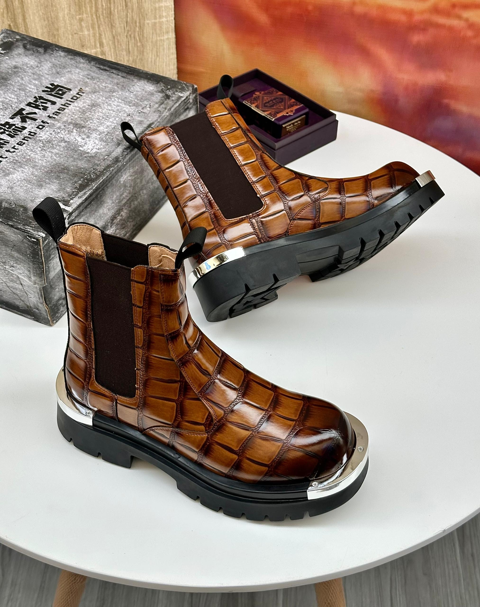 Brown crocodile-embossed Chelsea boots with metal toe cap and thick black lug sole on white table.