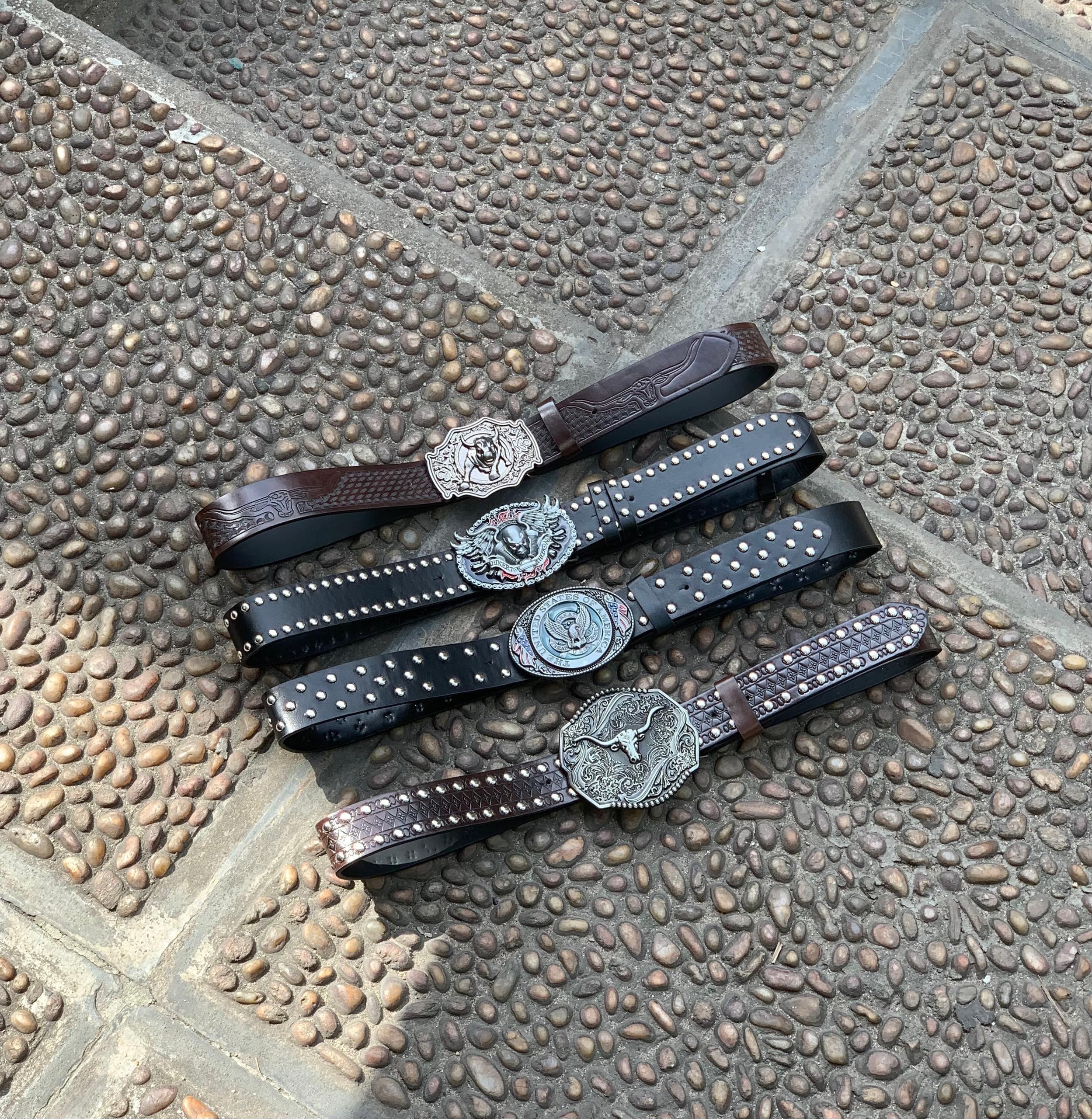 Collection of tooled leather belts with western themed buckles on cobblestone.