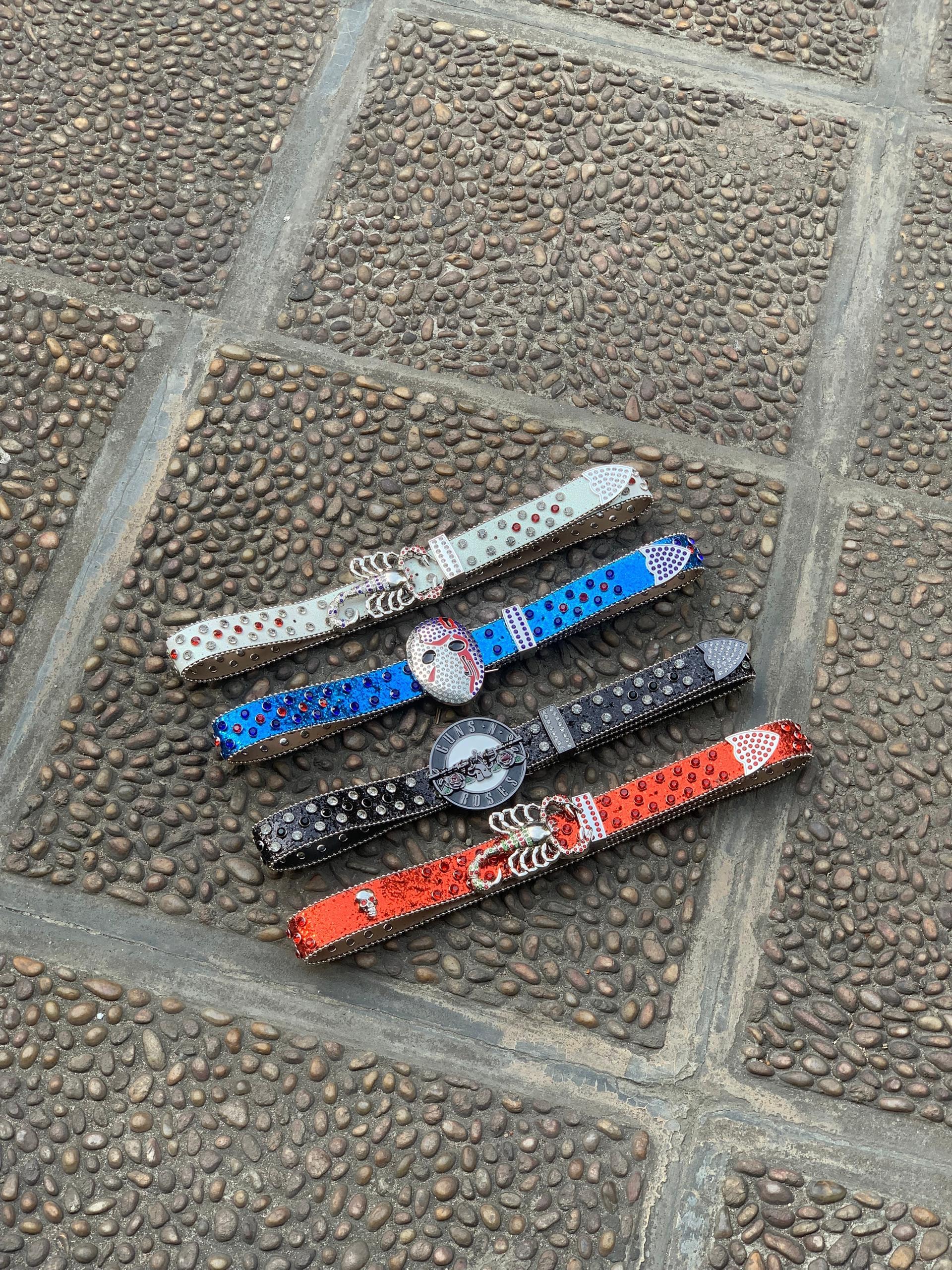 Four jeweled belts in white, blue, black and red with skull and Guns N Roses buckles on cobblestone.