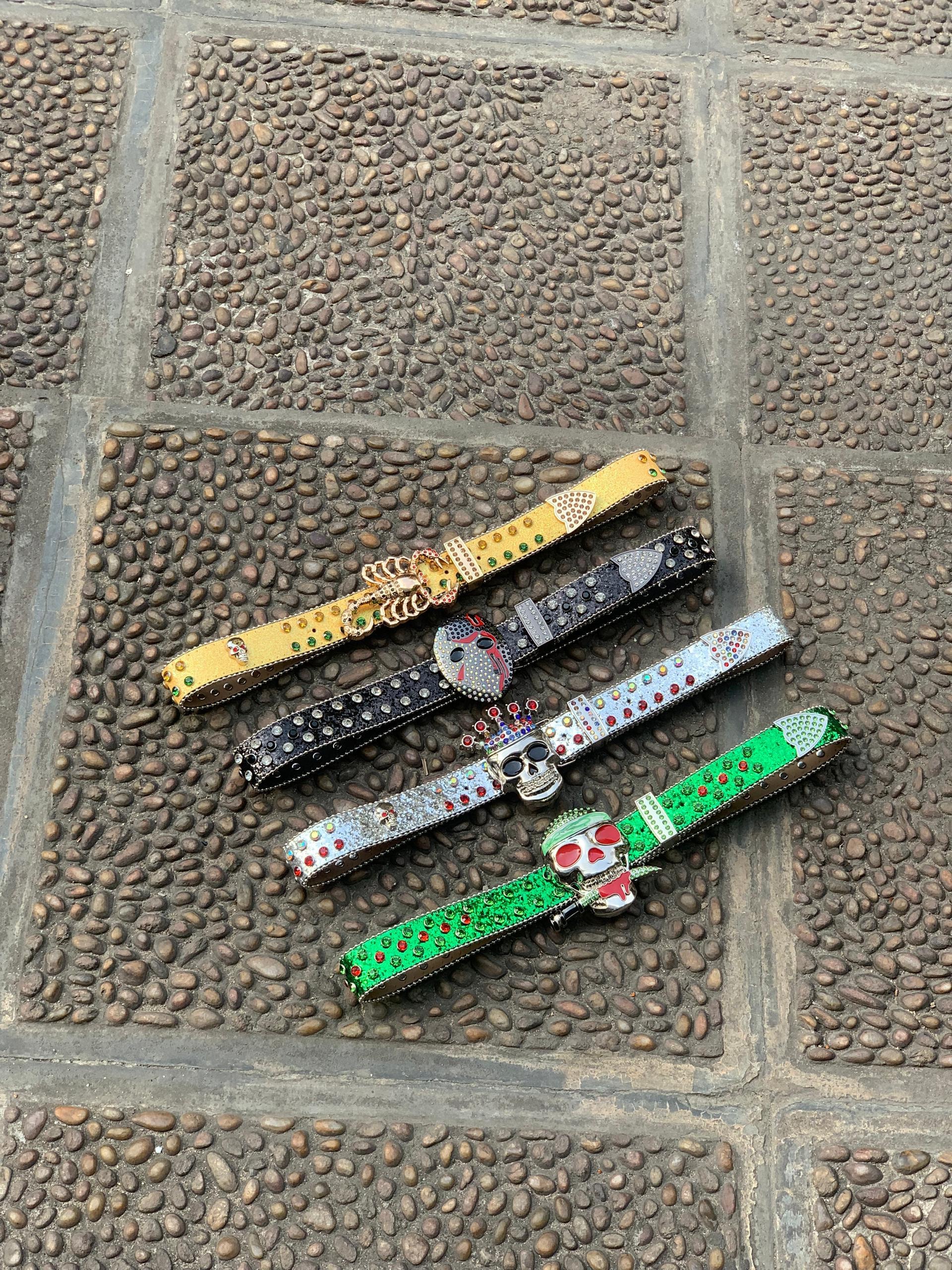 Four jeweled skull belts in yellow, black, silver and green with decorative buckles on cobblestone.