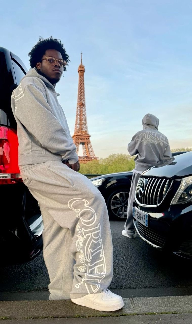 Man in gray Corteiz hoodie and joggers posing by car in front of Eiffel Tower.