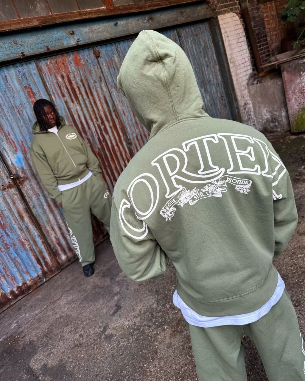 Model wearing olive green Corteiz 2-piece set from back showing large CORTEIZ graphic print.
