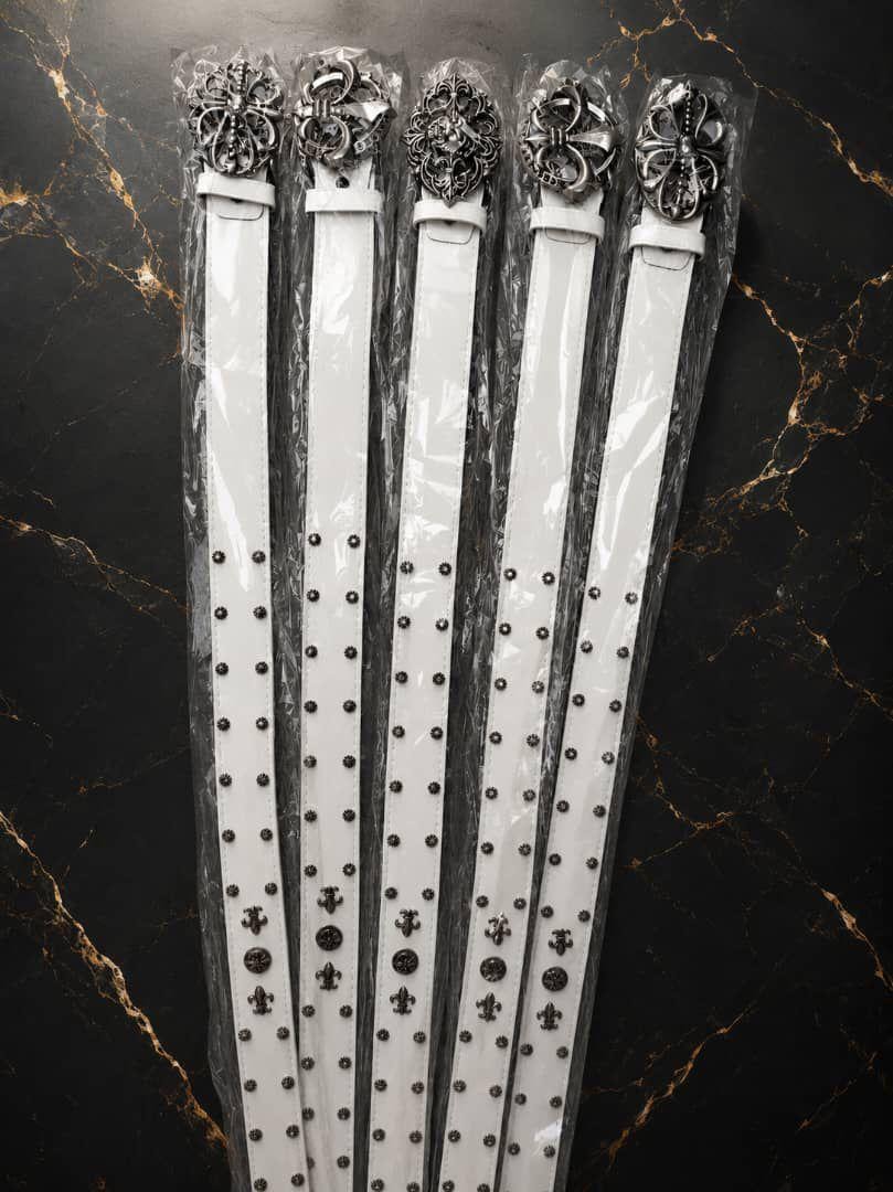 Five packaged designer belts with intricate silver buckles on marble background.