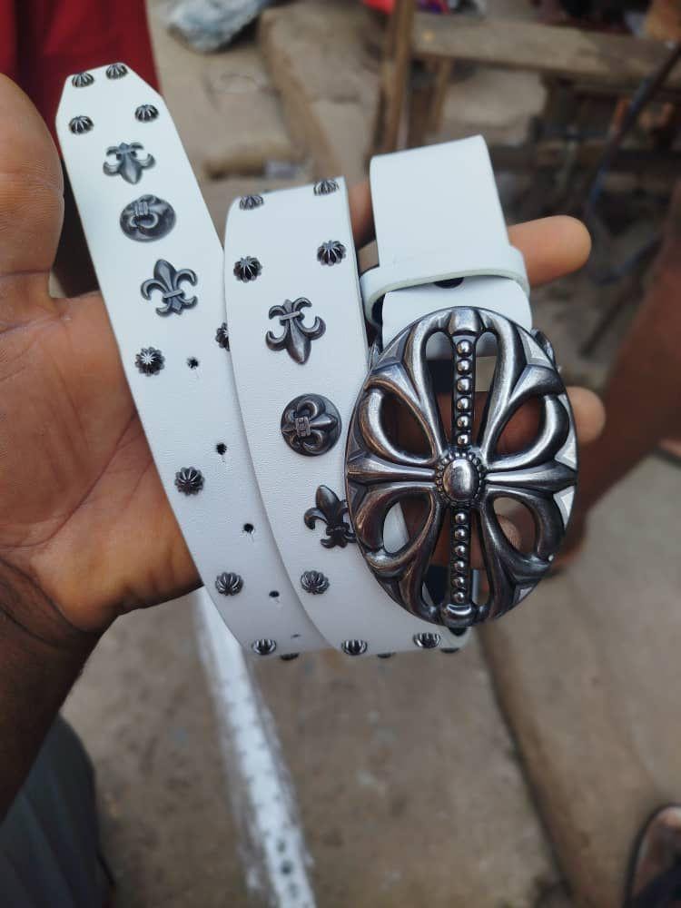 Hand holding white designer belt showing silver studs, emblems and large decorative buckle.