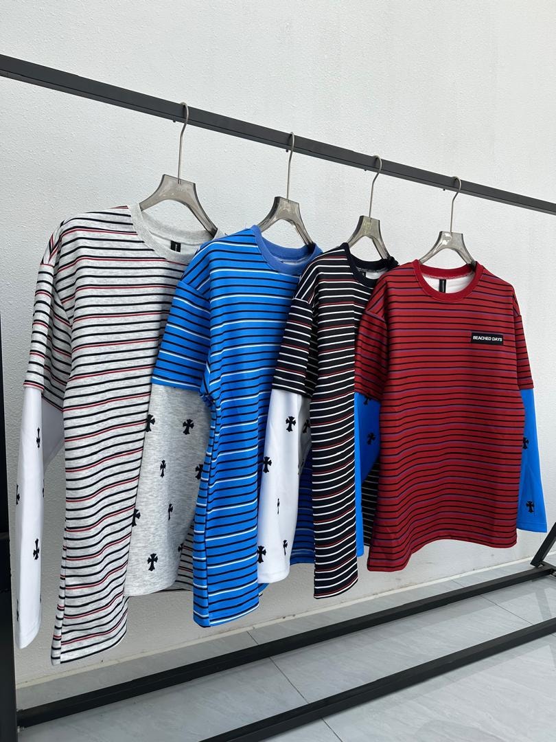 Four colorways of striped long sleeve t-shirts with cross patterns on sleeves hanging on rack.