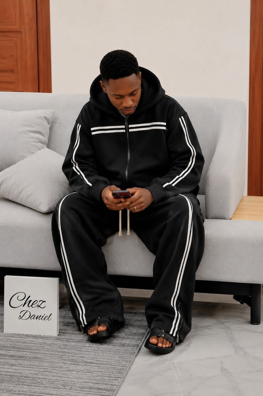 Man seated on couch wearing black zip-up striped hoodie and matching pants set.