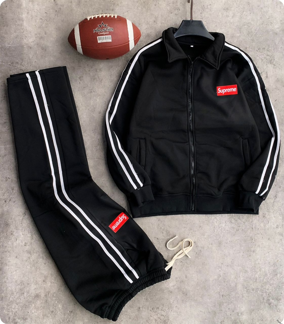 Black Supreme tracksuit set with zip jacket, striped pants, and football prop on gray background.