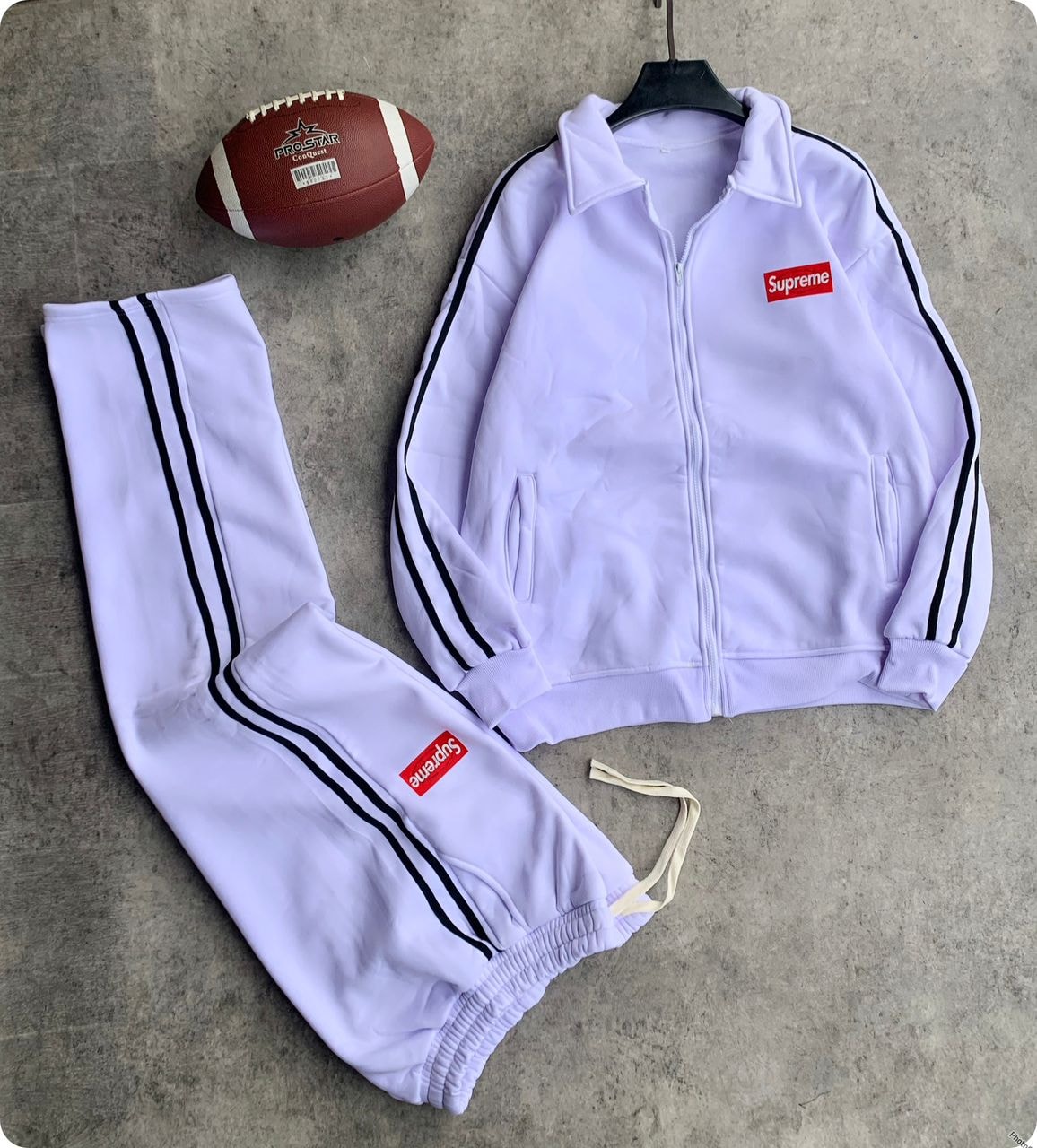 Purple Supreme tracksuit set with zip jacket, striped pants, and football prop on gray background.