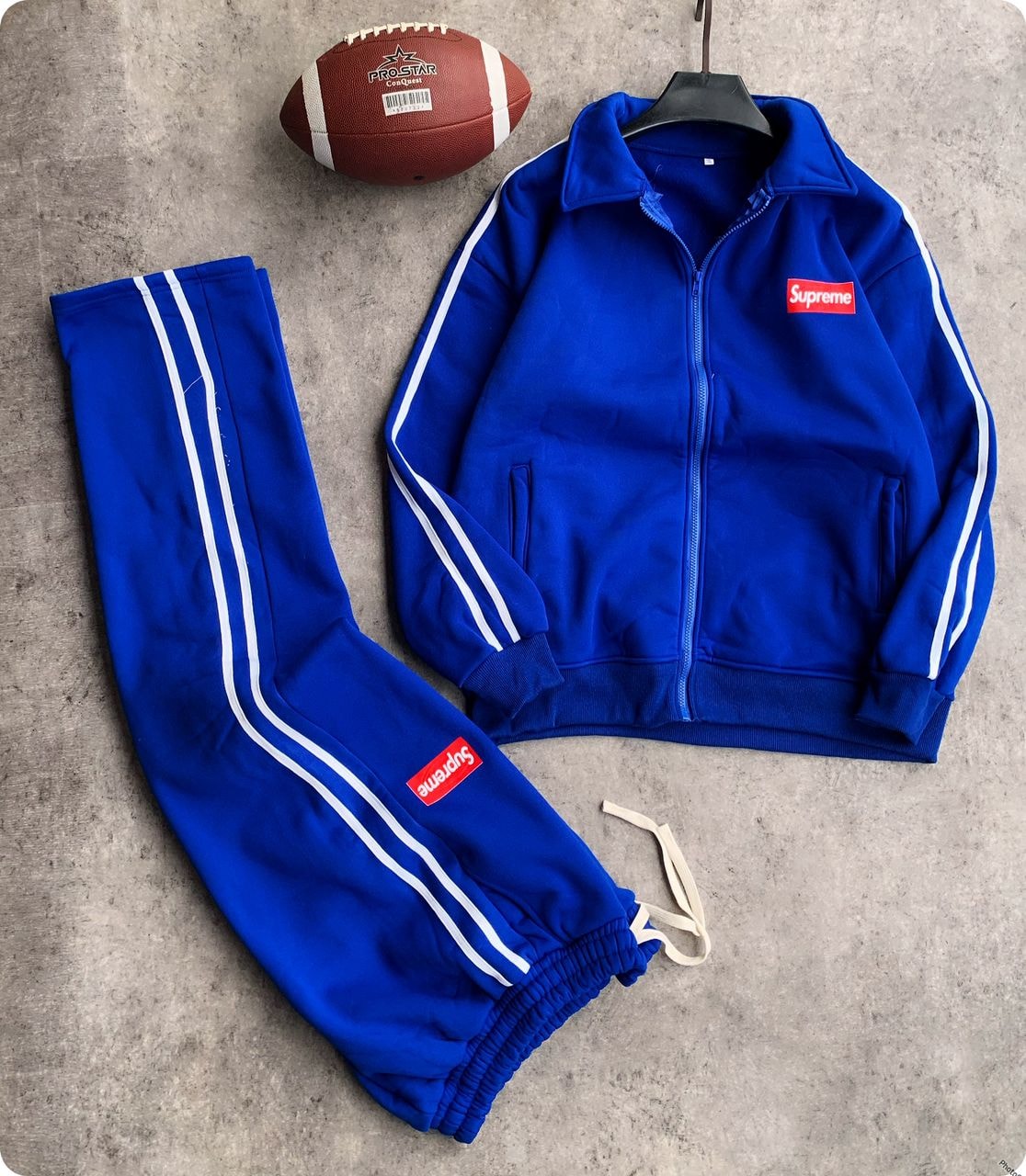 Blue Supreme tracksuit set with zip jacket, striped pants, and football prop on gray background.