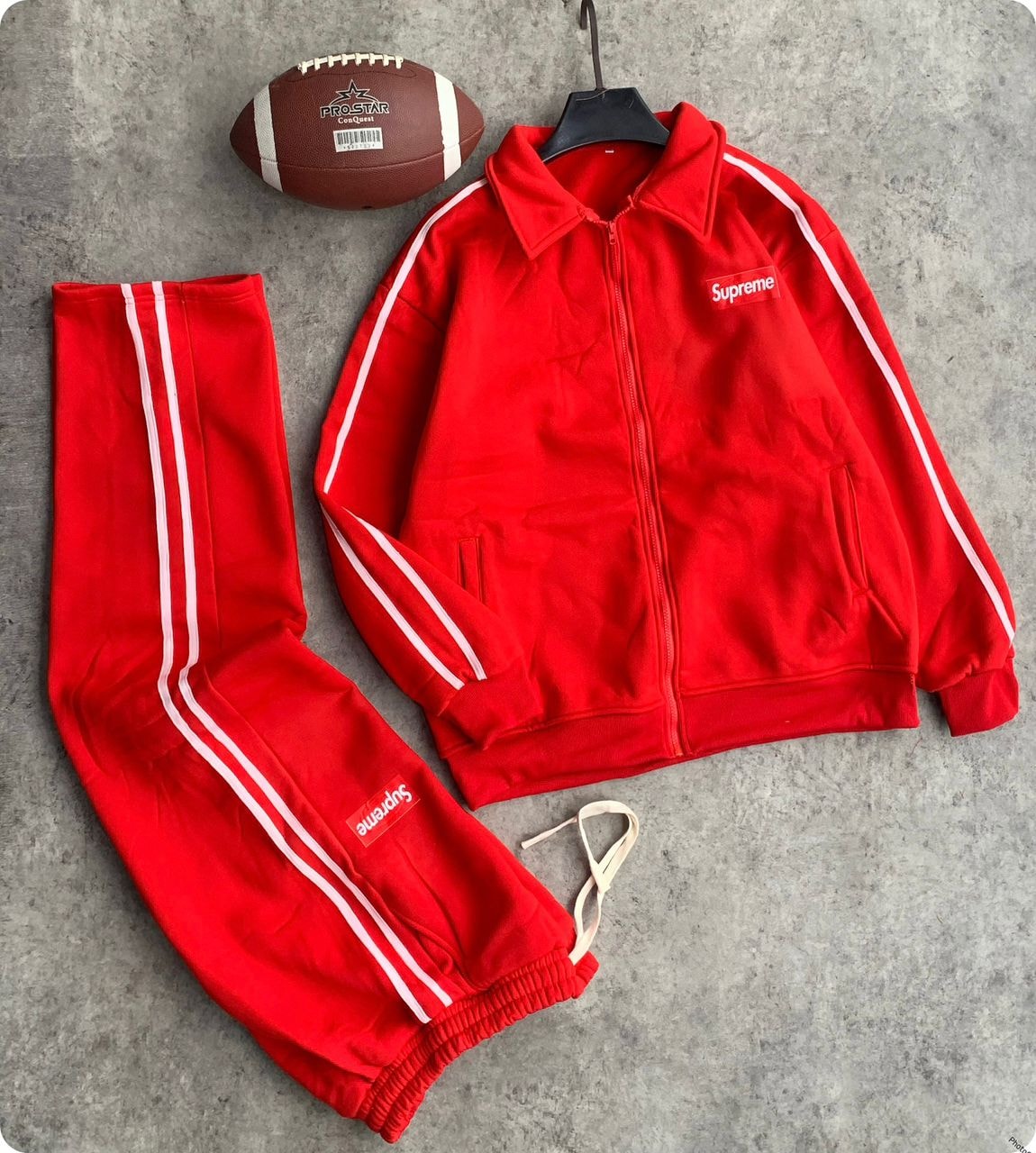 Red Supreme tracksuit set with zip jacket, striped pants, and football prop on gray background.