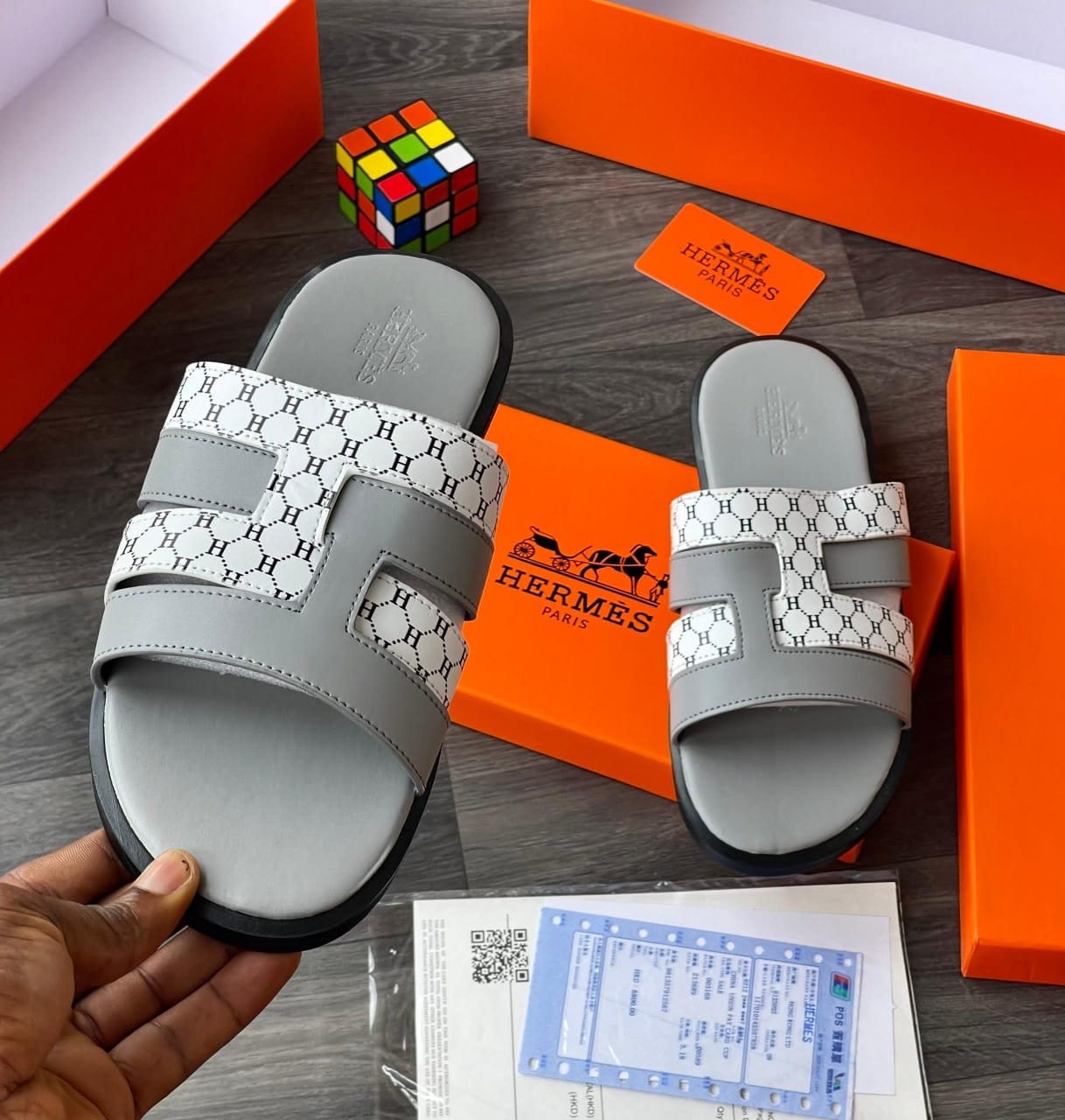 Grey Hermes monogram slides with H pattern held in hand alongside orange boxes and accessories.