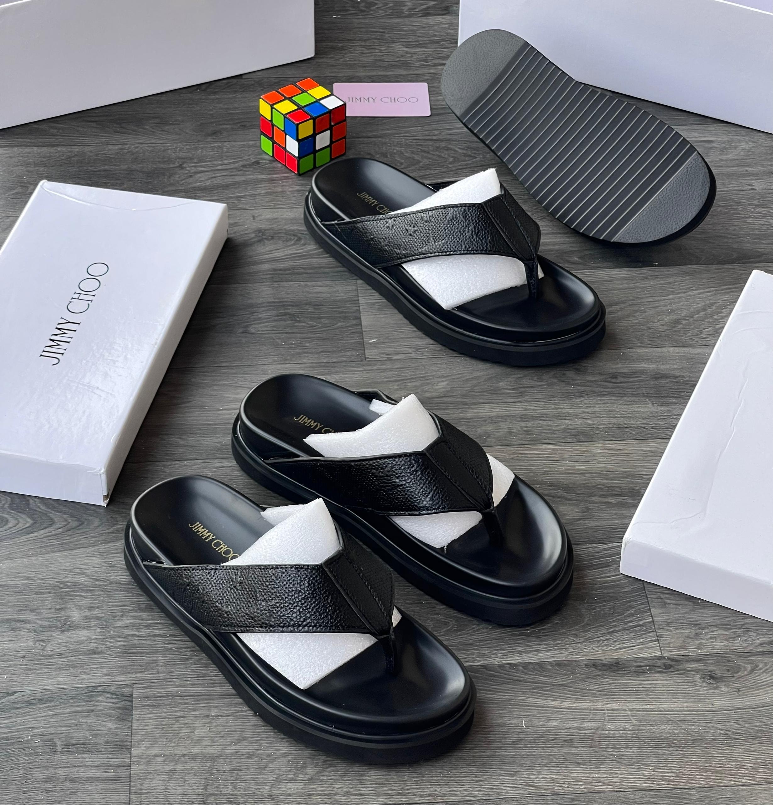 Black Jimmy Choo textured slides with white boxes, card and Rubik's cube on wooden floor.