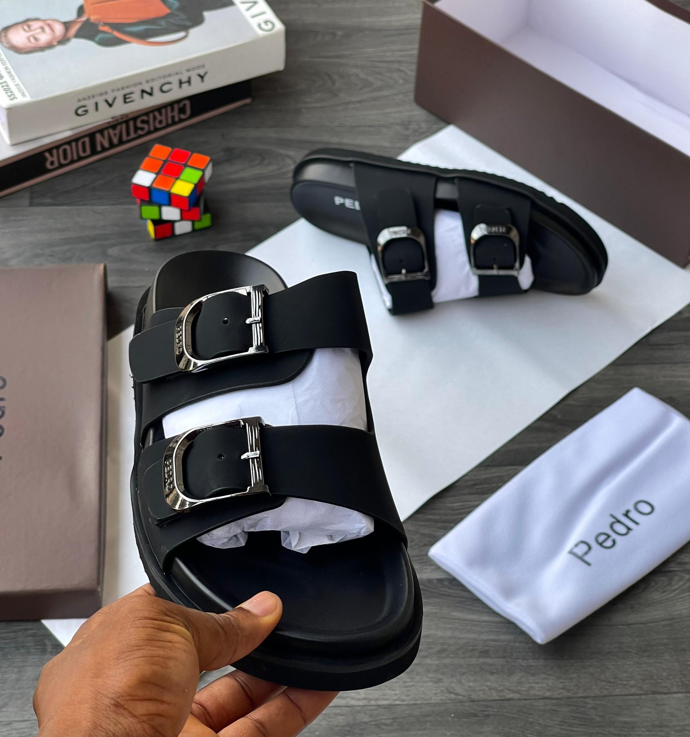 Black Pedro double buckle slides held in hand with boxes, books and Rubik's cube in background.