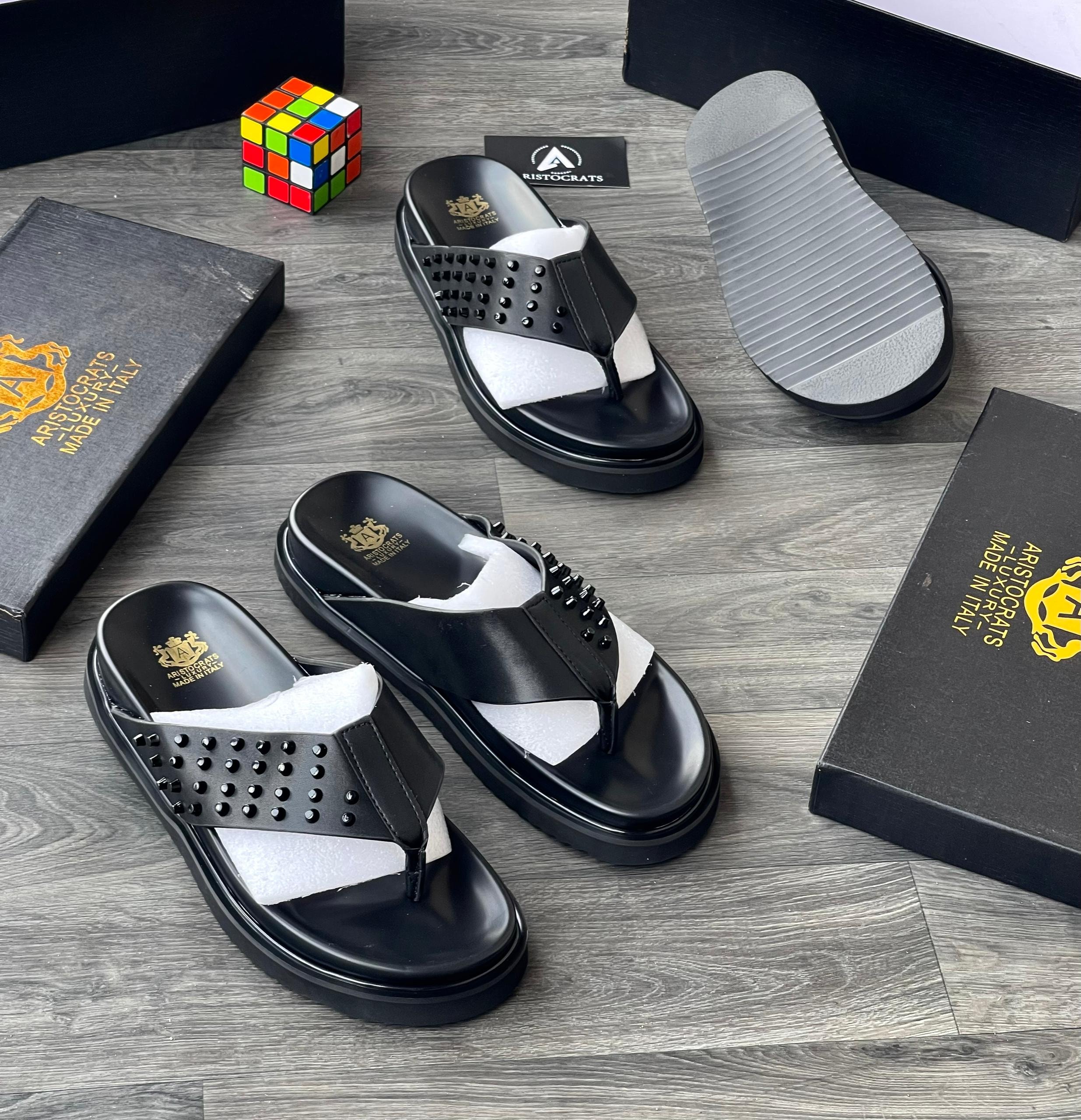 Black Aristocrats studded thong slides shown with black boxes, card and Rubik's cube.