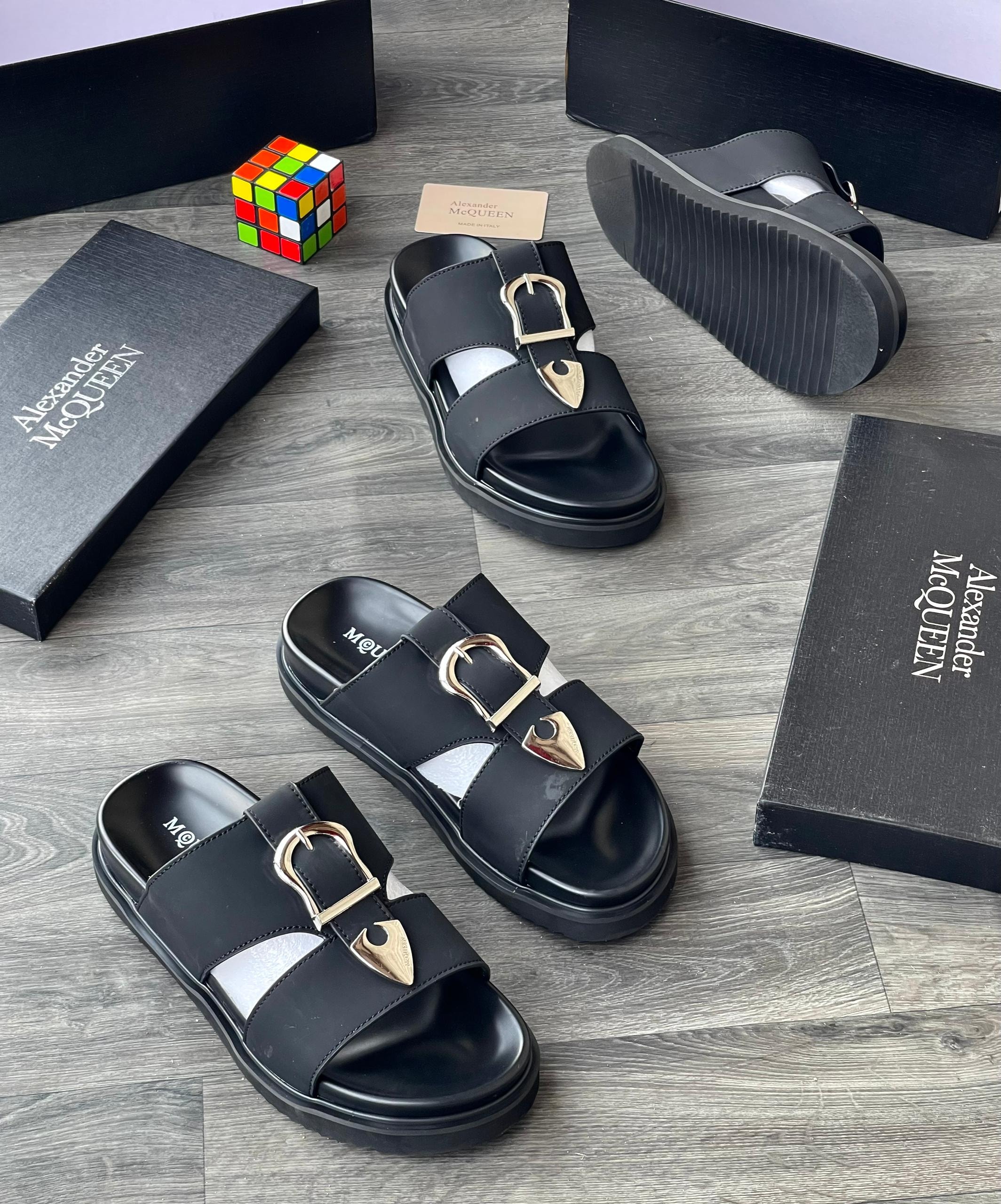 Black Alexander McQueen slides with gold buckle and arrow detail shown with black boxes and Rubik's cube.