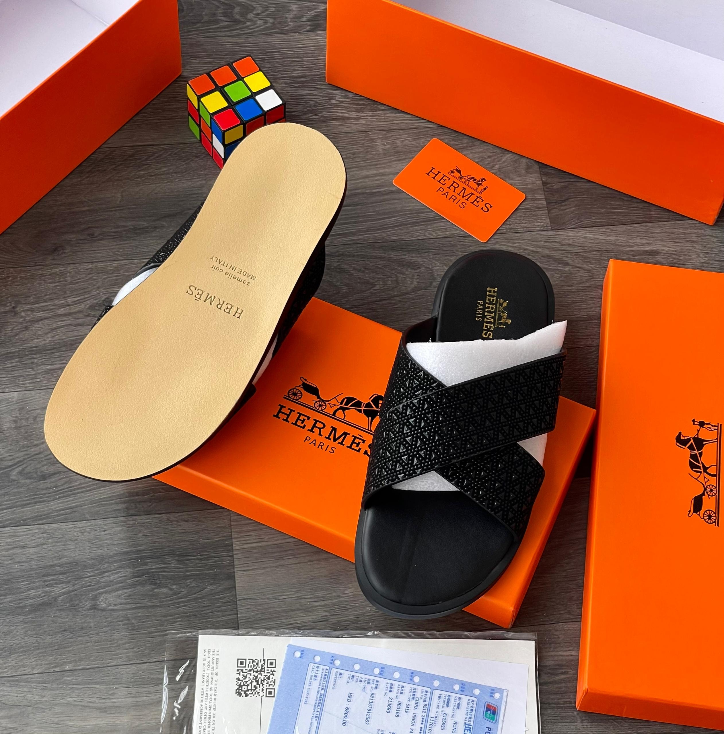 Black Hermes cross-strap slides with geometric pattern displayed with orange box, card and accessories.